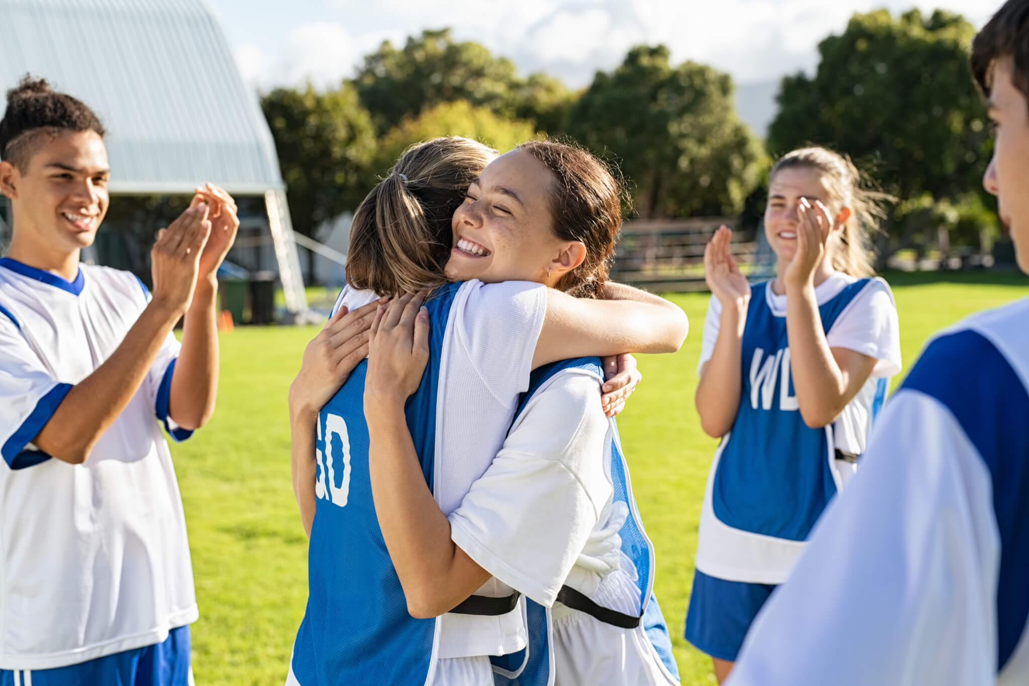 Two girls in sports kit hug in celebration while their team mates applaud. 