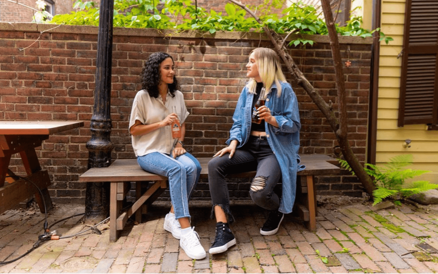 Two young women socialising on a bench with a drink each. 
