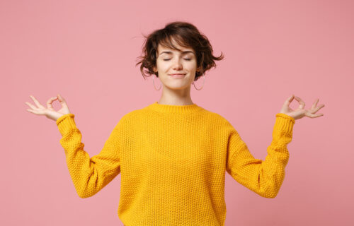 Young brunette woman girl in yellow sweater posing isolated on pastel pink background. People lifestyle concept. Mock up copy space. Hold hands in yoga gesture relaxing meditating keeping eyes closed